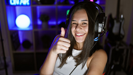 Young, beautiful hispanic woman streamer confidently gives a thumb-up, all smiles, in her game-filled gaming room. okay, it's game night, hey headset, waving that thumb in the dark home office.