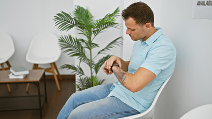 A pensive man with tattoos sits in a minimalist office lobby, checking his watch, evoking a sense...
