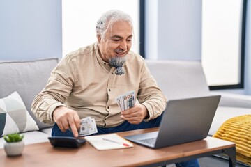 Middle age grey-haired man using laptop counting dollars at home