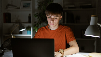 Caucasian teenage boy studying at night in a home office with a laptop, lamp, and books.