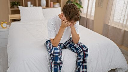 Young caucasian male teenager in pajamas sitting on bed in bedroom, looking stressed or sad.