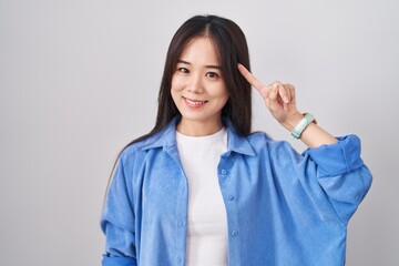 Young chinese woman standing over white background smiling pointing to head with one finger, great...