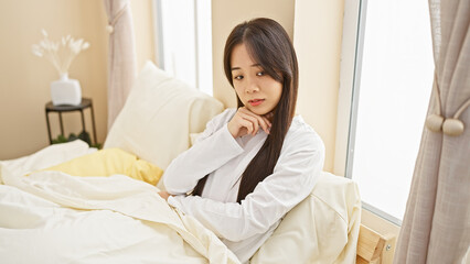 A young asian woman in a white shirt sitting thoughtfully on a bed in a well-lit bedroom interior