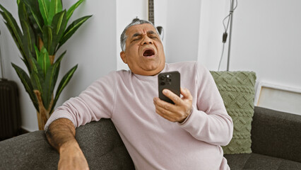 Mature man yawning with smartphone in a cozy living room, expressing fatigue or boredom.