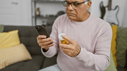 Middle-aged man in glasses reads medicine label at home, smartphone in hand, sitting on couch.