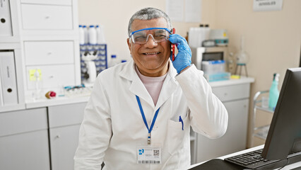 Smiling mature man in lab coat and safety goggles talking on phone in a laboratory setting