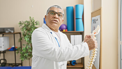Naklejka na ściany i meble Mature man therapist in white coat at rehab clinic interior examining a spine model