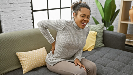 African american woman experiencing back pain while sitting on a grey sofa in a modern living room.