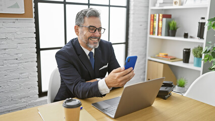Charming young hispanic man, grey-haired employee, engages in smiling video call, effortlessly...