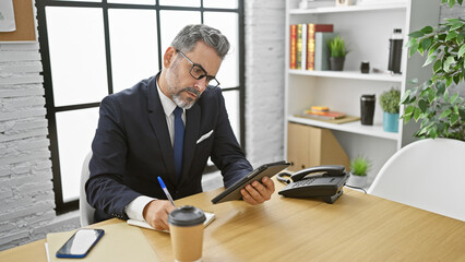 Attractive, grey-haired, young hispanic man, fully concentrated at work in his office, taking notes on touchpad, handsome professional with beard, glasses and suit sitting at table indoors - obrazy, fototapety, plakaty