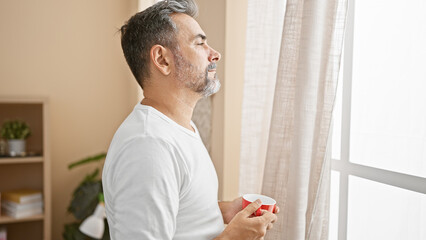 Serious young hispanic man, grey-haired & handsome, relaxed indoors at home. he's throwing a concentrated look through the window, clutching his morning coffee cup. - obrazy, fototapety, plakaty