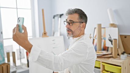 Handsome young hispanic, grey-haired male artist engrossed in a serious video call at indoor art studio, painting with paintbrush on canvas, learning and discussing art lessons.