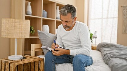 Young hispanic grey-haired man sitting on bed taking notes at bedroom