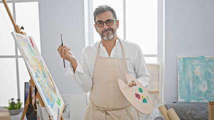 Confident young hispanic man, an artist with grey hair, joyfully stands smiling in an art studio,...