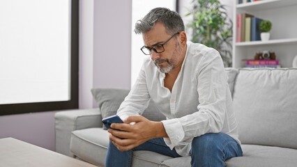 Young hispanic grey-haired man using smartphone sitting on sofa at home