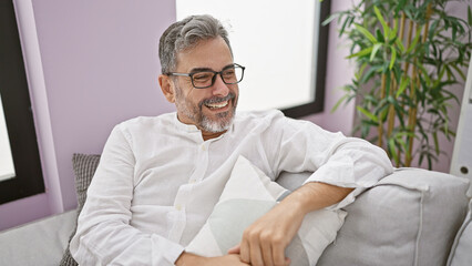 Cheerful, grey-haired hispanic man sits joyful and relaxed on living room sofa at home, smiling...