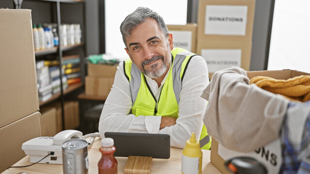 Beaming young hispanic man with grey hair, volunteering at a local charity center, confidently navigating the touchpad while sitting amidst donation boxes