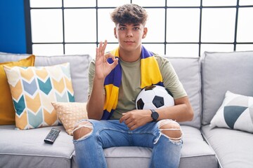 Hispanic teenager sitting on the sofa watching football match doing ok sign with fingers, smiling...