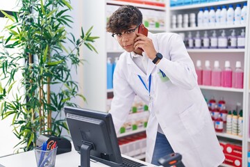 Young hispanic teenager pharmacist talking on smartphone using computer at pharmacy