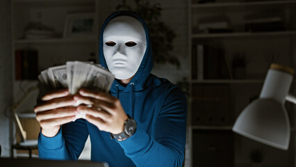 Masked man in hoodie holding us dollars at night in a dimly lit office, suggesting illicit...