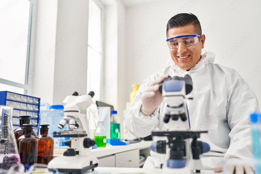 Wall mural Young latin man scientist wearing covid protection uniform using microscope at laboratory