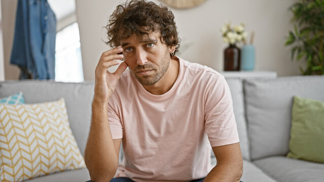 A thoughtful young hispanic man sits on a grey couch in a cozy living room, giving off a contemplative vibe.