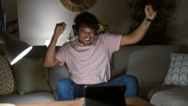 Excited young hispanic man with headphones celebrating victory at home on a comfortable sofa looking at a tablet screen.