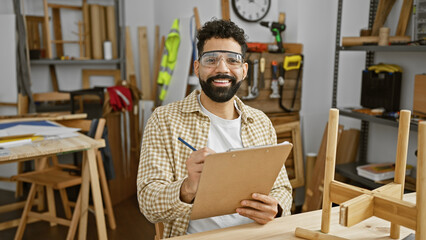 Handsome hispanic man with a beard takes notes in a carpentry workshop, wearing safety glasses