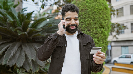 Smiling bearded man talking on phone and holding credit card in urban park