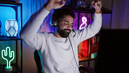 A joyful hispanic man with a beard celebrates in a gaming room with ambient lighting at night.