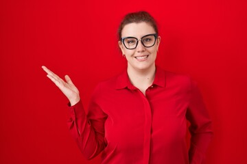 Young hispanic woman with red hair standing over red background smiling cheerful presenting and pointing with palm of hand looking at the camera.