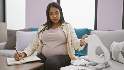 Serene young hispanic pregnant woman lovingly checking baby clothes and taking notes while...