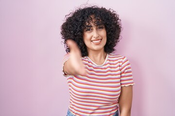 Young middle east woman standing over pink background smiling friendly offering handshake as...