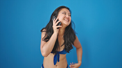 Fototapeta premium Young chinese woman tourist wearing bikini talking on smartphone over isolated blue background