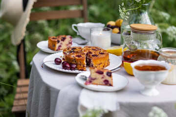 Tea time with sweet homemade cherry cake and herbal beverage in summer garden outdoors. Table covered with linen tablecloth, white porcelain cups. English style