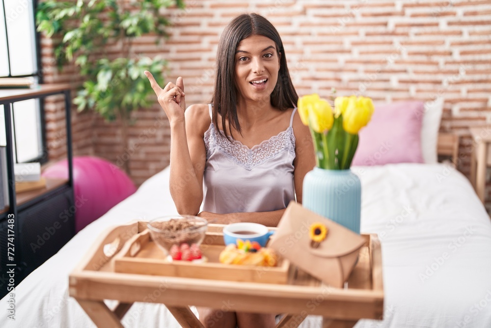 Wall mural Brunette young woman eating breakfast sitting on the bed smiling happy pointing with hand and finger to the side