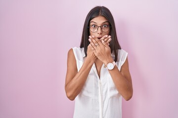 Brunette young woman standing over pink background wearing glasses shocked covering mouth with...