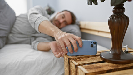 A middle-aged hispanic man with a beard reaching for a smartphone in his bedroom