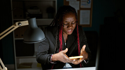 Exhausted african american business woman stressed out at work, texting problems away on smartphone in dark office room - obrazy, fototapety, plakaty