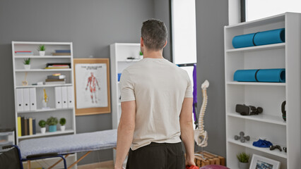 A young hispanic man wears casual attire in a modern rehabilitation clinic, viewed from behind with a focus on health.