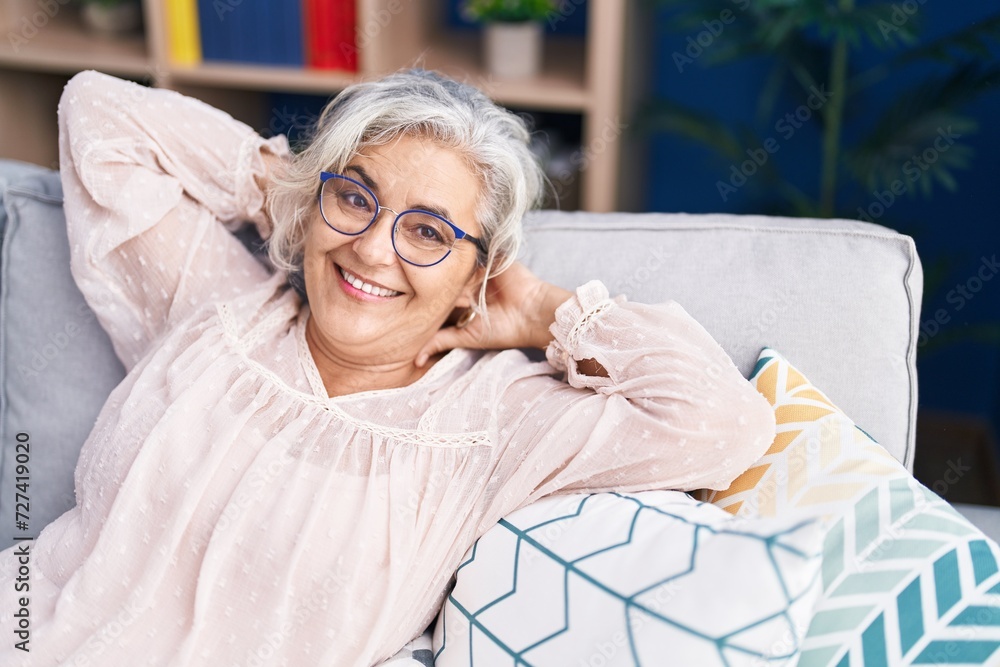 Wall mural middle age grey-haired woman relaxed with hands on head sitting on sofa at home