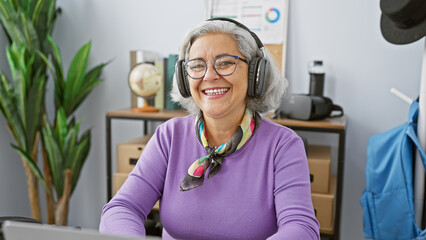 Smiling mature woman with headphones in modern office setting radiates positivity and...
