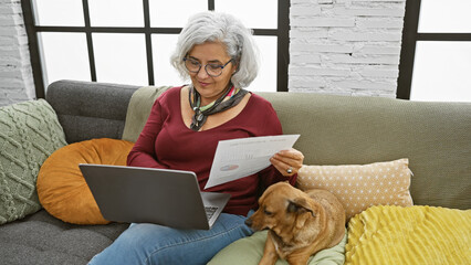 Mature woman reading paper with dog beside on couch indoors