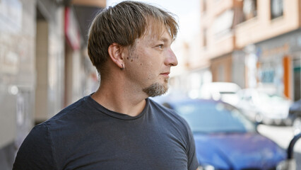 A handsome young adult man with a beard and blond hair stands outdoors on a city street, providing a contemporary urban feel.