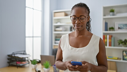 African american woman with glasses using smartphone in modern office setting. - obrazy, fototapety, plakaty