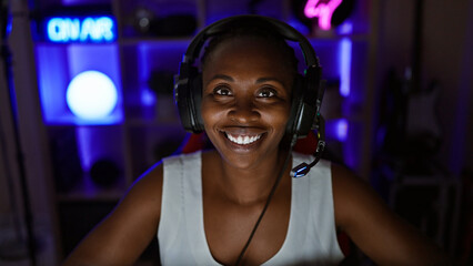A smiling young african woman wearing headphones in a vibrant gaming room at home, illuminated by...