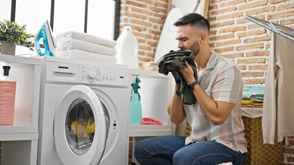Young hispanic man sitting on floor with serious face smelling dirty clothes at laundry room