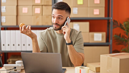 Young hispanic man ecommerce business worker talking on smartphone eating muffin at office