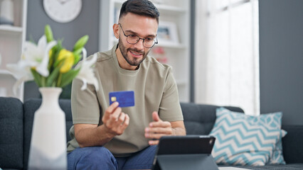 Young hispanic man shopping with touchpad and credit card sitting on sofa at home - obrazy, fototapety, plakaty