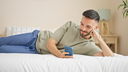 Fototapeta premium Young hispanic man using smartphone lying on bed at bedroom